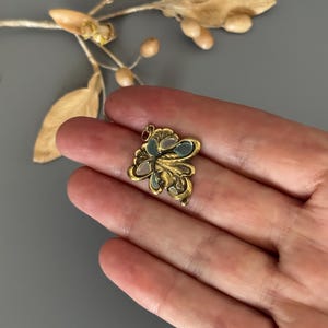 May include: A small, gold-colored pendant with an ornate floral design. The pendant features a central cluster of pale blue and white enamel, surrounded by detailed gold filigree. The piece is held in a hand against a gray background.