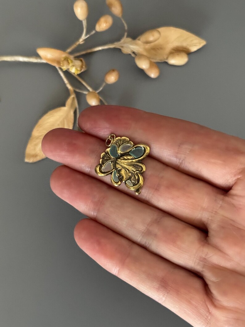 May include: A small, gold-colored pendant with an ornate floral design. The pendant features a central cluster of pale blue and white enamel, surrounded by detailed gold filigree. The piece is held in a hand against a gray background.