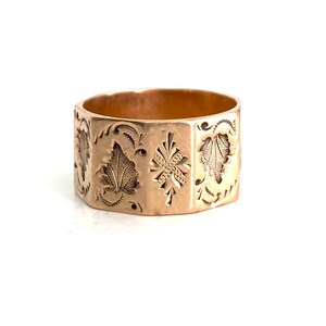 May include: A gold band ring with an intricate engraved design featuring leaves and a cross. The ring is made of a solid metal and has a polished finish.
