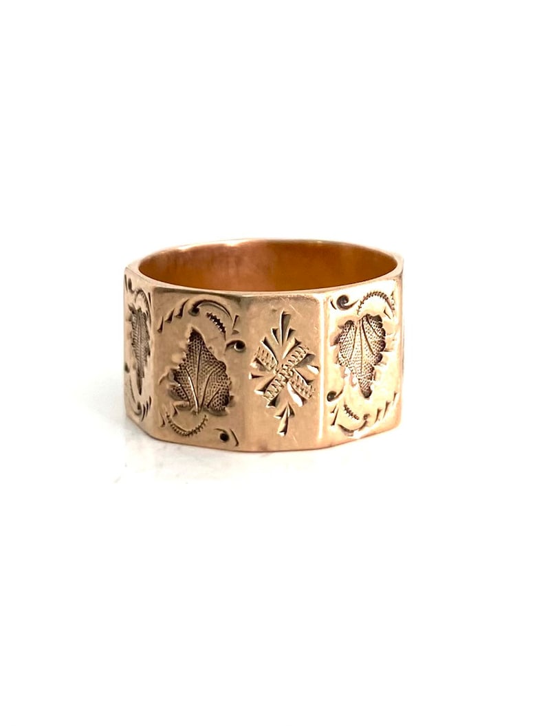 May include: A gold band ring with an intricate engraved design featuring leaves and a cross. The ring is made of a solid metal and has a polished finish.