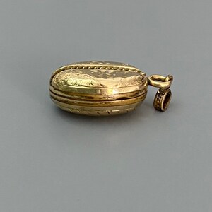 May include: A gold locket with an engraved design and a loop for a chain. The locket is closed and has a smooth, polished surface.