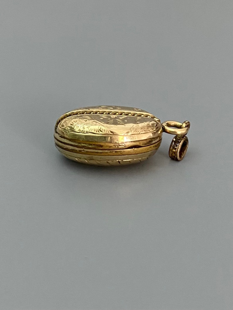 May include: A gold locket with an engraved design and a loop for a chain. The locket is closed and has a smooth, polished surface.