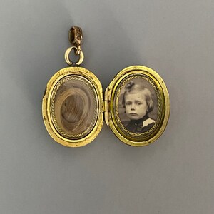 May include: Gold locket with a hinged lid. The locket is open, revealing a lock of hair in one side and a black and white photograph of a young person in the other side.