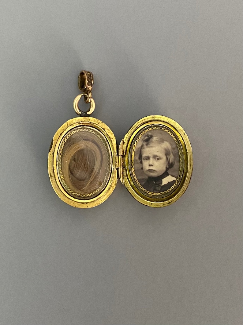 May include: Gold locket with a hinged lid. The locket is open, revealing a lock of hair in one side and a black and white photograph of a young person in the other side.