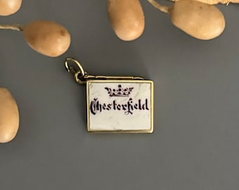 Può includere: Un piccolo medaglione dorato con sfondo in smalto bianco e una corona viola e la parola "Chesterfield" scritta in lettere viola.