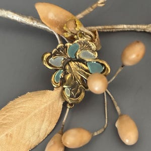 May include: Close-up of a vintage floral hair accessory. The piece features a gold-toned metal flower with teal and white enamel accents, a small face detail, and attached beige-colored leaves and berry-like beads. The accessory is set against a gray background.