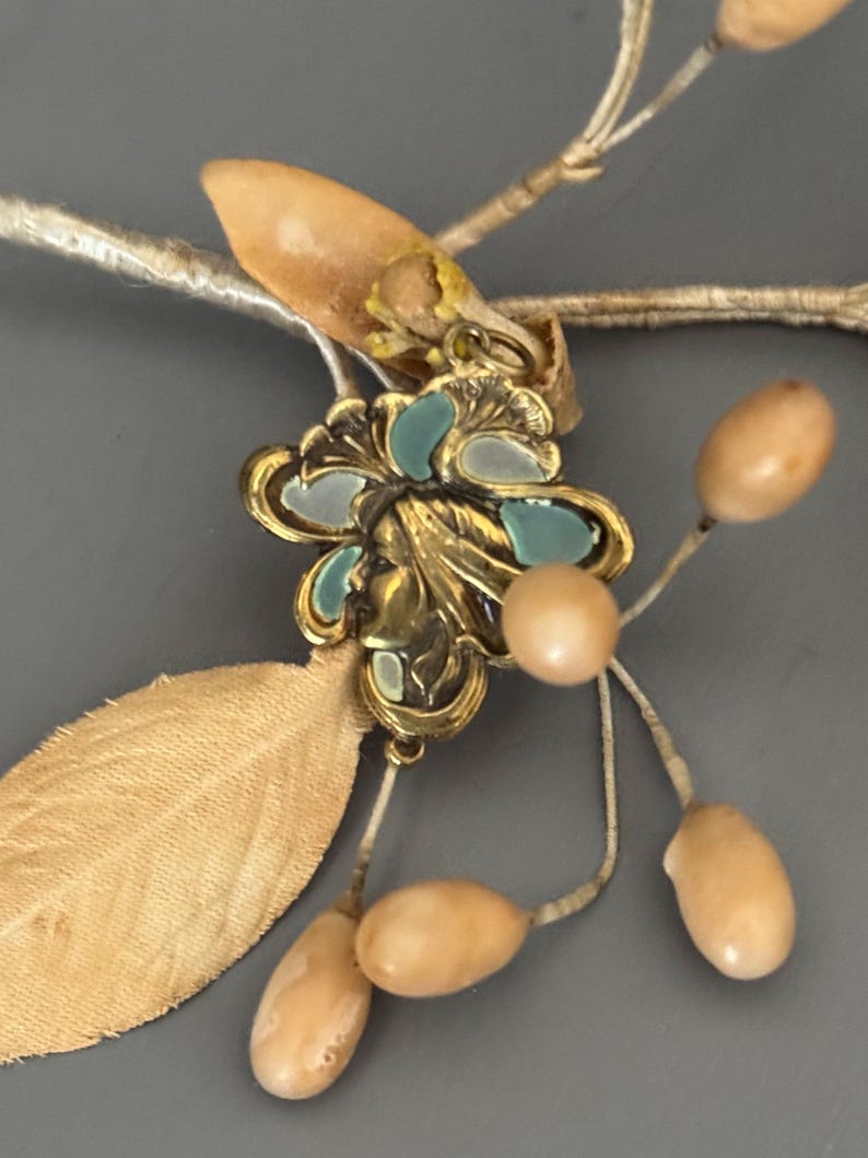 May include: Close-up of a vintage floral hair accessory. The piece features a gold-toned metal flower with teal and white enamel accents, a small face detail, and attached beige-colored leaves and berry-like beads. The accessory is set against a gray background.