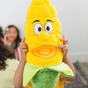 May include: A plush corn on the cob puppet with a smiling face, large eyes, and a green base. The puppet is yellow with a light orange mouth and is being held by a person wearing a yellow shirt with white polka dots.