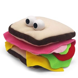Puede incluir: Un juguete de sándwich de peluche con grandes ojos saltones. El sándwich presenta capas de fieltro rosa, amarillo, verde y rojo, imitando el relleno. El pan es beige con bordes marrones.