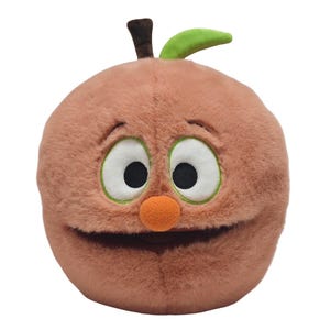 May include: A peach-coloured plush toy in the shape of an apple with a cartoon face. It has large eyes with green outlines, a small orange nose, and a smiling mouth. It features a brown stem and a green leaf.