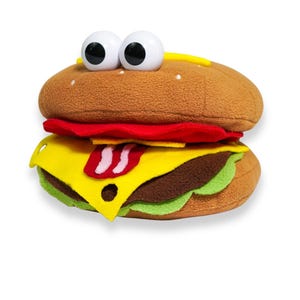 May include: A plush burger toy with googly eyes. The burger features a brown bun, red tomato, yellow cheese, a brown patty, and green lettuce. The toy has a playful design, perfect for kids.
