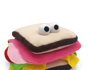 May include: A plush toy shaped like a sandwich with googly eyes. The sandwich is made of brown, pink, yellow, and green felt.