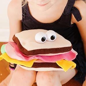 Op de afbeelding: Een pluchen sandwichspeelgoed met lichtbruin brood, een donkerbruine korst en kleurrijke vullingen van roze, geel en groen. De sandwich heeft grote, wiebelende ogen. Het wordt vastgehouden door een persoon die een zwarte jurk draagt.