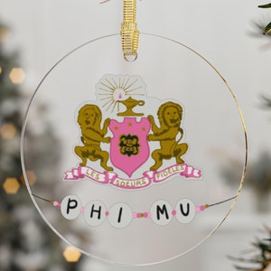 May include: Clear, round Christmas ornament with a gold ribbon. The ornament features a pink crest with gold lions and the text "LES SOEURS FIDELES". Below, the letters "PHI MU" are spelled out with white and pink beads.