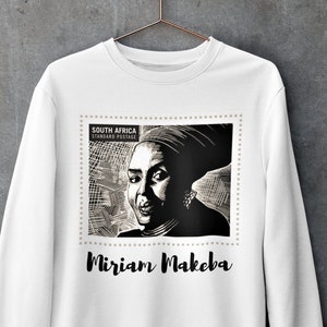 Peut inclure: Sweat-shirt blanc avec une image en noir et blanc de Miriam Makeba sur un timbre postal sud-africain. Le texte "SOUTH AFRICA STANDARD POSTAGE" et "Miriam Makeba" sont imprimés sur le sweat-shirt.