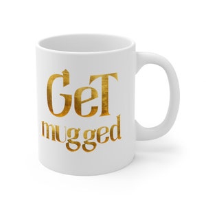 Puede incluir: Taza de caf&eacute; de cer&aacute;mica blanca con un dise&ntilde;o met&aacute;lico dorado que dice "Get mugged".