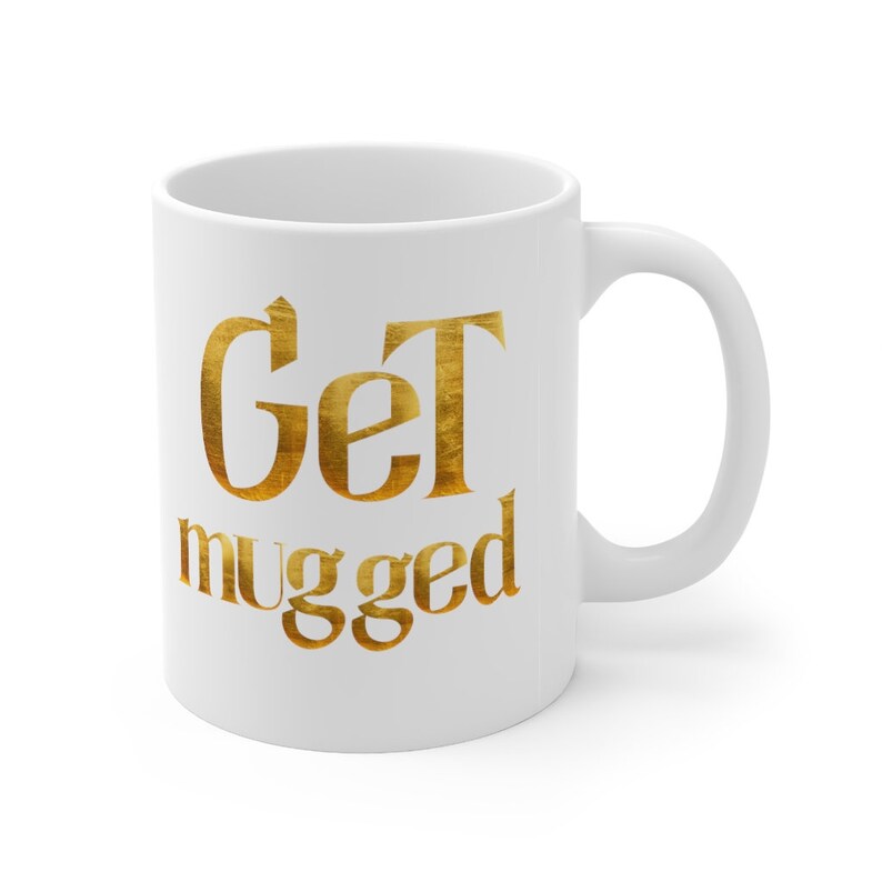Puede incluir: Taza de caf&eacute; de cer&aacute;mica blanca con un dise&ntilde;o met&aacute;lico dorado que dice "Get mugged".