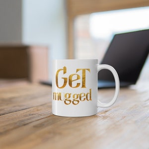 Puede incluir: Taza de cer&aacute;mica blanca con una inscripci&oacute;n dorada que dice "Get mugged".