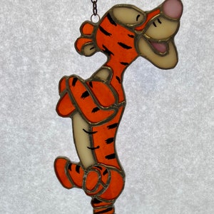 Könnte beinhalten: Ein hängender Glasbild-Anhänger mit Tigger aus Winnie Puuh. Tigger ist orange und schwarz mit einem weißen Gesicht und einer rosa Nase. Er ist in einer verspielten Pose mit seinem eingerollten Schwanz dargestellt.