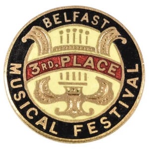May include: A round, enamel pin with the text "BELFAST MUSICAL FESTIVAL" in black lettering. The pin features a cream-colored center with a red banner that reads "3rd PLACE". Gold and black accents complete the design.