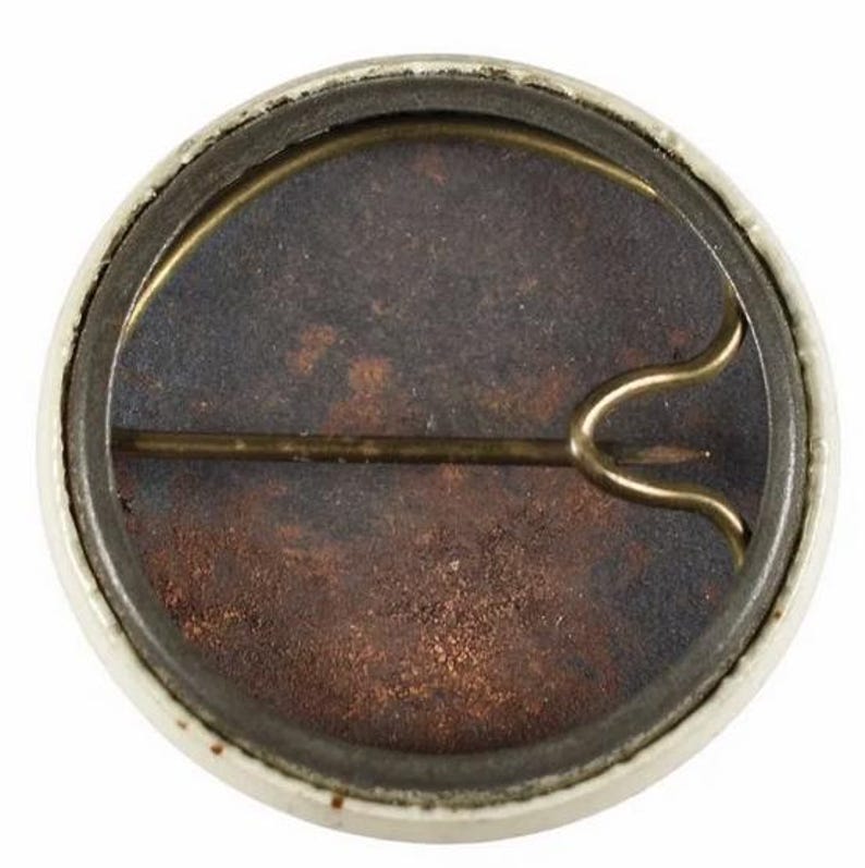 K&ouml;nnte beinhalten: Ein runder, alter Knopf mit wei&szlig;em Rand und rostbraunem Innenraum. Die R&uuml;ckseite zeigt eine Messingnadel und einen Verschluss zum Befestigen. Das Design des Knopfes deutet darauf hin, dass es sich um einen &auml;lteren Artikel handelt, m&ouml;glicherweise ein Sammlerst&uuml;ck oder ein Erinnerungsst&uuml;ck.