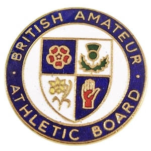 Può includere: Una spilla rotonda smaltata con bordo blu e la scritta "BRITISH AMATEUR ATHLETIC BOARD". Il centro presenta uno scudo bianco con quattro quadranti, ognuno contenente un emblema diverso in rosso, verde e giallo.