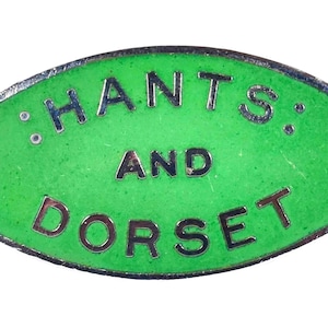 May include: An oval green enamel badge with the words "HANTS AND DORSET" in silver lettering. The badge has a silver border and a gold-coloured pin clasp. A vintage collectible item.