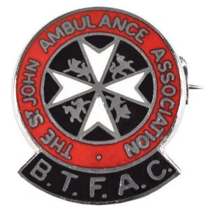 May include: A round pin with a red border, featuring the text "ST JOHN AMBULANCE ASSOCIATION" and "B.T.F.A.C.". The pin has a white Maltese cross at the center, surrounded by a black background and silver accents.