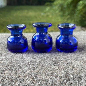 May include: Three small, cobalt blue glass vases with a rounded body and a flared rim. The vases are displayed on a gray surface, with a blurred green background. The vases are similar in shape and size, with a glossy finish.