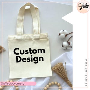 May include: A white tote bag with black handles and the text "Custom Design" printed on the front. The bag is sitting on a white surface with a wooden bead garland and a small bunch of cotton.
