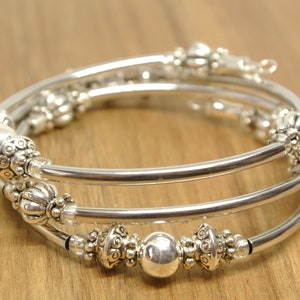 May include: A silver-toned, multi-strand bracelet with decorative beads. The bracelet features three parallel, smooth bands with ornate, textured beads and clear crystal accents. The beads have a floral design. The bracelet is shown on a wooden surface.