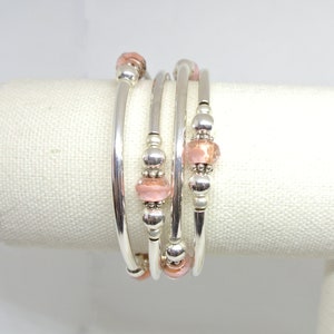 May include: A set of four silver-toned spiral bracelets. Each bracelet features a series of small, round silver beads and larger, irregular pink beads. The bracelets are displayed on a white, textured surface.