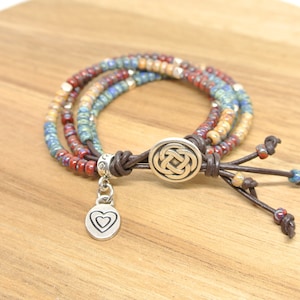 May include: A multi-strand bracelet with brown leather cord, silver heart charm, and a silver Celtic knot button closure. The bracelet features colorful glass beads in shades of red, blue, and beige.