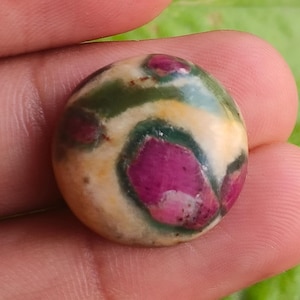 Könnte beinhalten: Ein runder, polierter Edelstein mit cremefarbener Basis, akzentuiert mit dunkelgrünen und leuchtend magenta Mustern. Der Stein wird in einer Hand gehalten, mit grünem Laub im Hintergrund. Der Edelstein hat einen Durchmesser von etwa 2,5 cm.