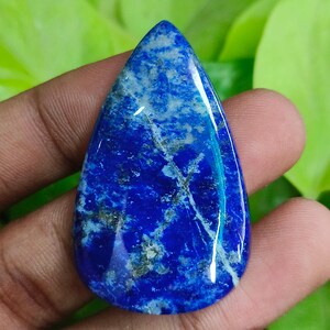 Könnte beinhalten: Ein tropfenförmiger Lapis-Lazuli-Edelstein. Der polierte Stein zeigt eine tiefblaue Farbe mit weißen Streifen und goldenen Flecken. Der Edelstein wird vor einem Hintergrund aus grünem Laub gehalten.