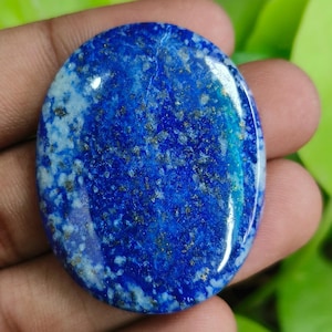 Könnte beinhalten: Ovaler Lapis Lazuli-Edelstein. Der Stein hat eine tiefblaue Farbe mit goldenen und weißen Flecken. Die polierte Oberfläche reflektiert das Licht und hebt die natürlichen Muster und Texturen hervor. Der Edelstein wird in einer Hand gehalten.