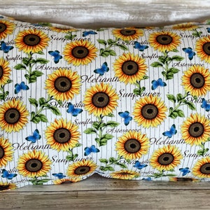 May include: White pillow with a sunflower pattern. The pillow has blue butterflies and the words "Helianthus", "Sunflower", "Asteraceae", and "Delitha" printed on it.