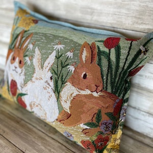 May include: A rectangular pillow with a green background featuring a brown rabbit and two white rabbits in a field of flowers. The pillow has a light blue border.