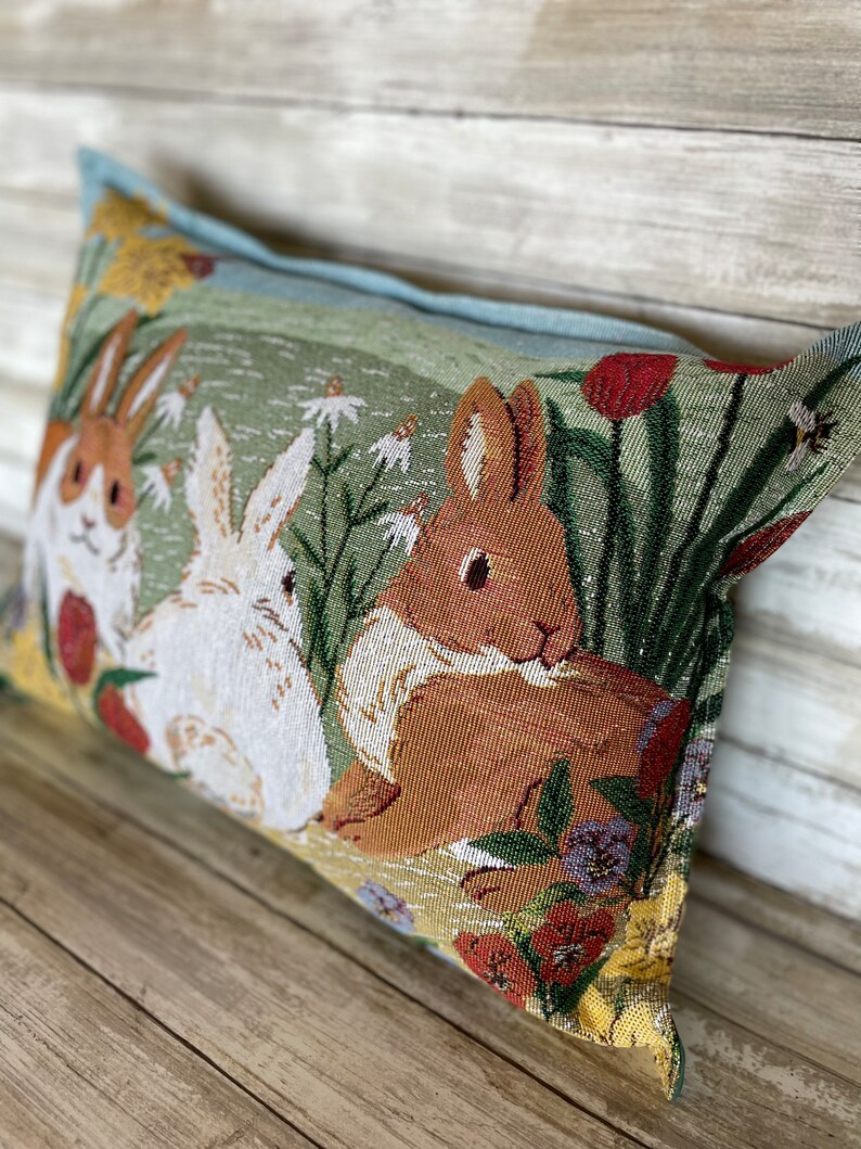 May include: A rectangular pillow with a green background featuring a brown rabbit and two white rabbits in a field of flowers. The pillow has a light blue border.