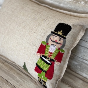 May include: A beige pillow with an embroidered nutcracker holding a green drum. The nutcracker is wearing a red and green uniform with gold accents and a black hat.