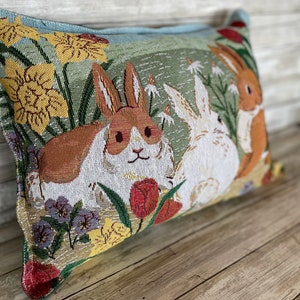 May include: A decorative pillow with a tapestry design featuring two rabbits in a field of flowers. The rabbits are brown and white, and the flowers are yellow, red, and purple.