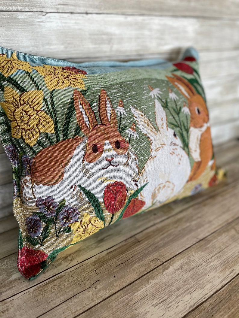 May include: A decorative pillow with a tapestry design featuring two rabbits in a field of flowers. The rabbits are brown and white, and the flowers are yellow, red, and purple.