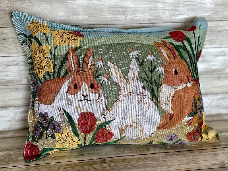 May include: A decorative pillow with a tapestry design featuring three rabbits in a field of flowers. The rabbits are brown and white, and the flowers are yellow, red, and purple.