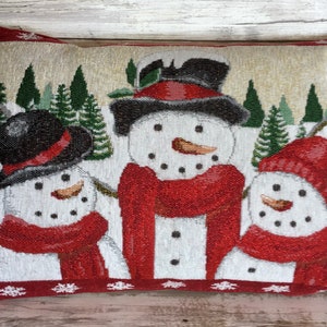 May include: A decorative throw pillow with a winter scene featuring three snowmen wearing red scarves and hats. The snowmen are standing in front of a snowy forest with green pine trees.