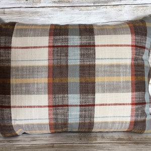 May include: Rectangular decorative pillow with a plaid pattern. The pillow features a brown, cream, gray, and light blue color scheme. The plaid design includes vertical and horizontal lines, creating a classic and cozy aesthetic. The pillow is made of a woven fabric.