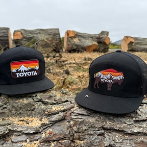 May include: Two black baseball caps with mesh backs. The left cap features a rectangular patch with a mountain range, sunset, and the word TOYOTA. The right cap features a bison-shaped patch with a mountain range, sunset, and the word TOYOTA.