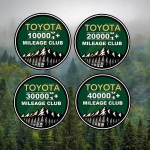 Puede incluir: Cuatro pegatinas circulares verdes y doradas con el texto "TOYOTA" y "MILEAGE CLUB". Cada pegatina presenta un número de kilometraje diferente: 10.000+, 20.000+, 30.000+ y 40.000+. Las pegatinas se colocan sobre un fondo borroso de árboles verdes.