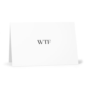 Può includere: Un biglietto bianco piegato con le lettere "WTF" stampate in nero, centrate sul davanti. Il biglietto è leggermente angolato, mostrando il suo bordo piegato e proiettando un'ombra morbida.