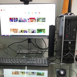 May include: A desktop computer setup featuring an HP monitor with a webcam mounted on top, a black tower PC unit with a vertical antenna, a black Logitech keyboard, and a black Logitech mouse. The monitor displays a Firefox browser window.