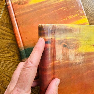 May include: Two hardback books with orange, yellow and red sunset-like covers. The books are stacked on top of each other.
